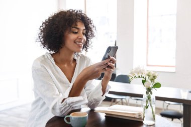 Gülümseyen Afrikalı güzel bir kadın kafeteryada dinleniyor, cep telefonu kullanıyor.