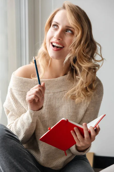 窓の近くにノートを書いている家の中で笑顔の若い女性の写真 — ストック写真