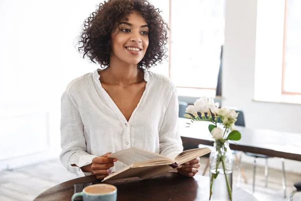 Lovely young african woman reading book while sitting at the cafe indoors drinking coffee