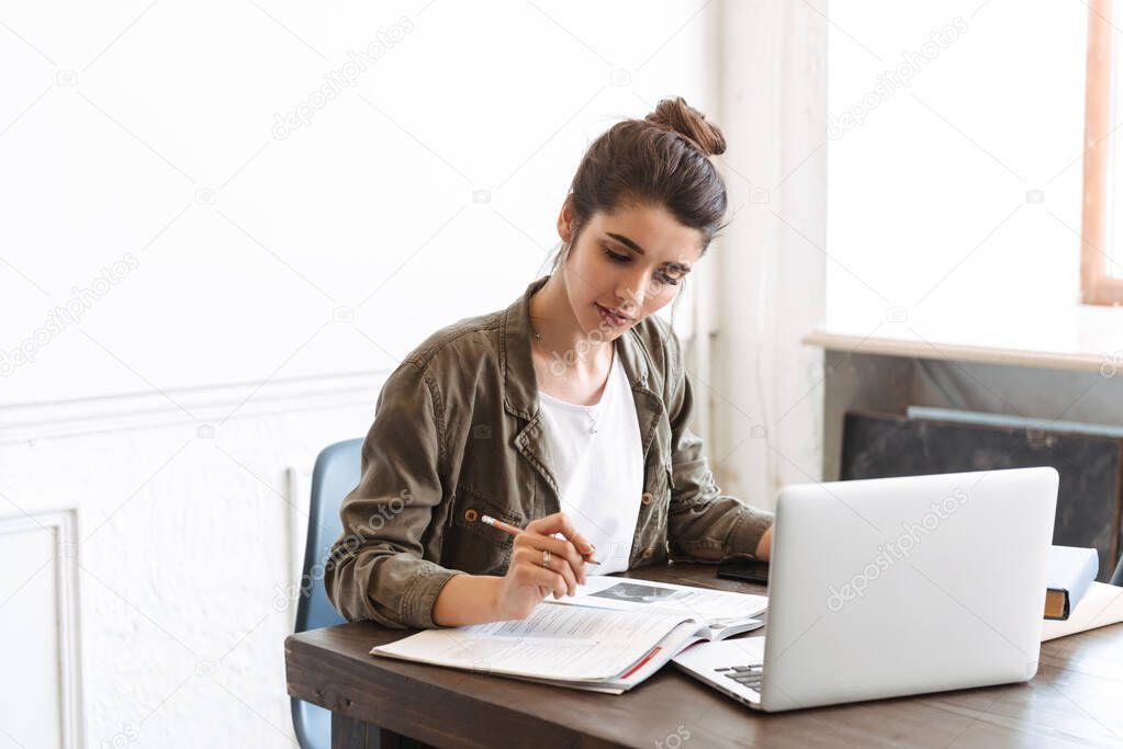 Image of a beautiful young concentrated woman using laptop computer indoors writing notes in notebook.