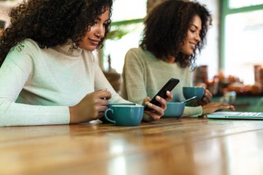 İki neşeli Afrikalı kız arkadaş kafede oturmuş kahve içiyor, cep telefonu ve dizüstü bilgisayar kullanıyorlar.