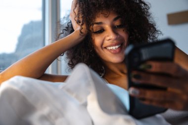 Evde cep telefonu kullanan neşeli, Afrikalı bir kadının görüntüsü yatakta yatıyor..