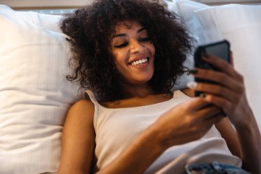 Evde cep telefonu kullanan neşeli, genç Afrikalı bir kadının fotoğrafı yatakta yatıyor..