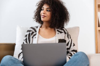 Evde dizüstü bilgisayar kullanan güzel ve memnun bir Afrikalı kadın resmi.