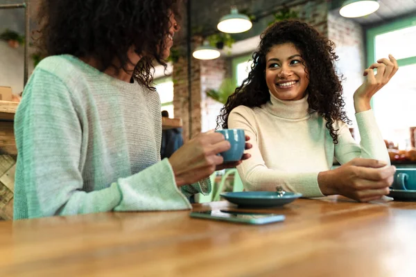 两个快乐的年轻非洲姑娘坐在咖啡馆边喝咖啡边聊天 — 图库照片