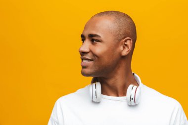 Çekici, güler yüzlü, rahat, sarı arka planda dikilmiş, kablosuz kulaklıkla müzik dinleyen Afrikalı genç bir adamın portresi.