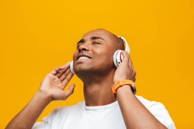 Çekici, güler yüzlü, rahat, sarı arka planda dikilmiş, kablosuz kulaklıkla müzik dinleyen Afrikalı genç bir adamın portresi.