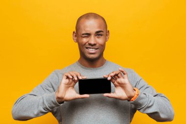 Çekici, gülümseyen, kendinden emin, sarı arka planda duran, boş ekranlı cep telefonunu gösteren Afrikalı genç bir adamın portresi.