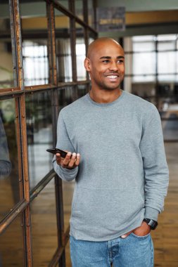 Ofiste çalışırken cep telefonu tutan ve sweatshirt giyen genç, kel, Afro-Amerikan bir adamın resmi.