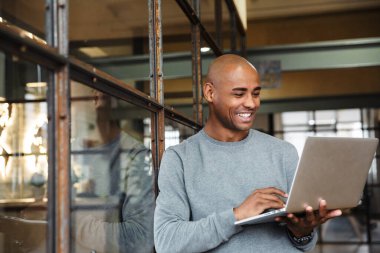 Ofiste çalışırken kucağında dizüstü bilgisayarla gezen yakışıklı, kel, Afro-Amerikan bir adamın resmi.