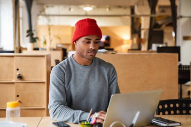 Genç, çekici, Afro-Amerikan bir adamın ofiste çalışırken dizüstü bilgisayar kullanarak şapka takması.