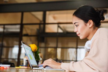 Ofiste dizüstü bilgisayarla çalışırken kahve içip not alan Asyalı güzel bir kadın resmi.