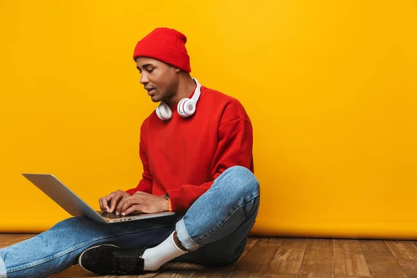一个有魅力 随和的年轻非洲男子的画像 他坐在黄色背景的地板上 用手提电脑 — 图库照片
