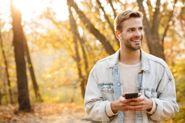 Parkta yürürken gülümseyen ve akıllı telefon kullanan kulaklıklı yakışıklı beyaz adam.