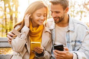 Sonbahar parkında bankta oturan neşeli genç çift cep telefonu kullanıyor.