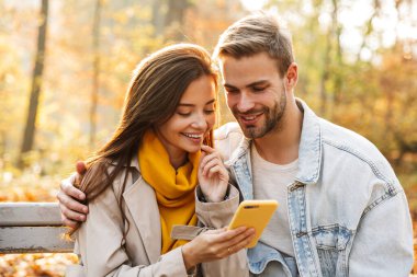 Sonbahar parkında bankta oturan neşeli genç çift cep telefonu kullanıyor.