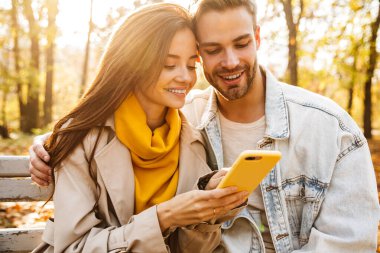 Sonbahar parkında bankta oturan neşeli genç çift cep telefonu kullanıyor.