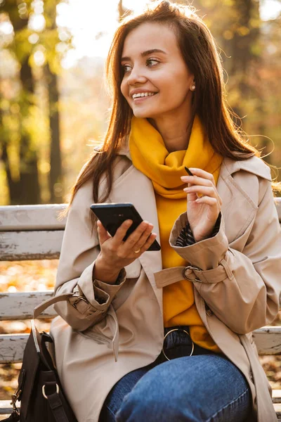 公園のベンチに座って 携帯電話を使って 秋のコートを着て長い髪の若い女性を笑顔 — ストック写真
