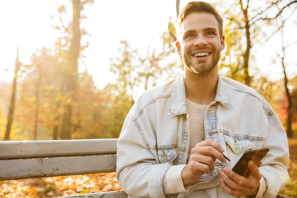 Hezký Usměvavý Mladý Muž Sedí Lavičce Podzimním Parku Používá Mobilní — Stock fotografie