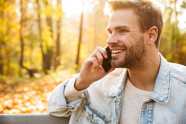 Hezký Usměvavý Mladý Muž Sedí Lavičce Podzimním Parku Mluví Mobilním — Stock fotografie