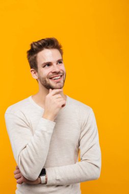 Çekici, mutlu, düşünceli, pulslu giyen genç adam sarı arka planda izole edilmiş, başka tarafa bakıyor.