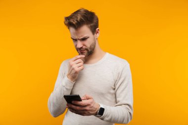 Çekici, kafası karışık genç adam sarı arka planda izole edilmiş, cep telefonu kullanıyor.