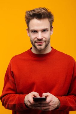Çekici mutlu genç adam sarı arka plan üzerinde izole edilmiş, cep telefonu kullanarak ayakta duruyor.