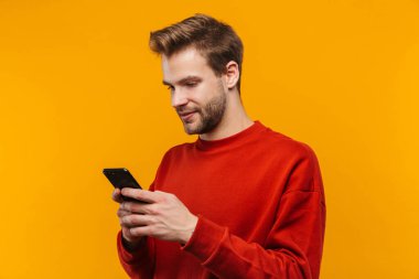 Pulmer giyen çekici genç adam sarı arka planda izole edilmiş, cep telefonu kullanıyor.