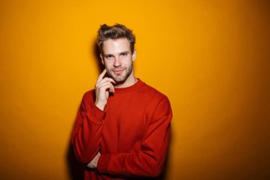 Çekici, düşünceli, gülümseyen, pulslu giyinen genç adam sarı arka planda tek başına duruyor.