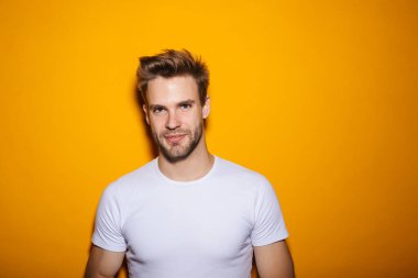 Çekici, gülümseyen, tişörtlü genç adam sarı arka planda dikilip kameraya bakıyor.