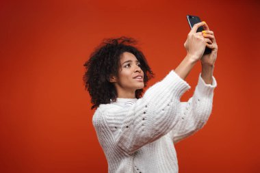 Gülümseyen çekici genç bir kadın kırmızı arka planda tek başına duruyor, selfie çekiyor.