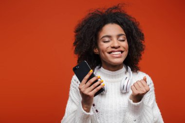 Çekici, gülümseyen, süveter giyen, kırmızı arka planda cep telefonu tutan Afrikalı bir kadın.