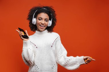 Neşeli, çekici, kablosuz kulaklık takan Afrikalı bir kadın kırmızı arka planda tek başına duruyor, cep telefonu tutuyor, dans ediyor.