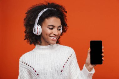 Kırmızı arka planda izole bir şekilde duran kablosuz kulaklık takan neşeli, çekici, genç Afrikalı kadın boş ekran cep telefonu gösteriyor.