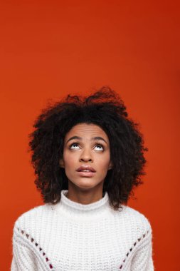 Düşünceli, genç bir Afrikalı kadın, günlük kıyafetler giyip kırmızı arka planda izole bir şekilde dikiliyor.