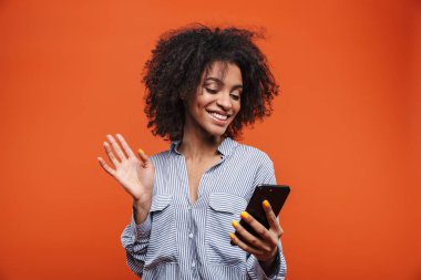 Kırmızı arka planda izole edilmiş cep telefonu kullanan gülümseyen Afrikalı genç bir kadın.