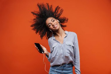 Gülümseyen, güzel bir Afrikalı kadın, günlük kıyafetler giyip kırmızı arka planda izole bir şekilde duruyor, kulaklıkla ve cep telefonuyla müzik dinliyor, dans ediyor.