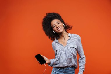 Gülümseyen, güzel bir Afrikalı kadın, günlük kıyafetler giyip kırmızı arka planda izole bir şekilde duruyor, kulaklıkla ve cep telefonuyla müzik dinliyor, dans ediyor.