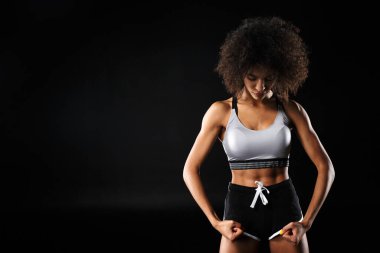 Afro-Amerikalı genç bir kadının spor kıyafetleri içinde poz verirken ve bisepslerini siyah arka planda izole bir şekilde gösterirken görüntüsü