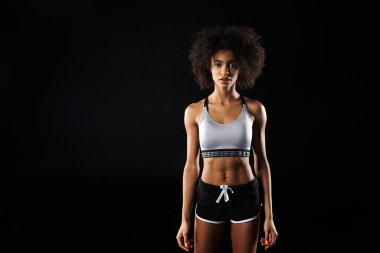 Baştan çıkarıcı Afro-Amerikan kadın spor kıyafetleri içinde siyah arka planda poz verip kameraya bakarken görüntüsü