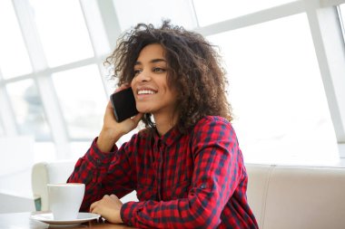 Ekose gömlek giyen, kahve içen ve kafede cep telefonuyla konuşan güzel Afro-Amerikan kadının portresi.