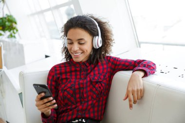 Ekose gömlek giyen ve kafede kulaklık ve cep telefonuyla müzik dinleyen Afrikalı Amerikalı genç bir kadının portresi.