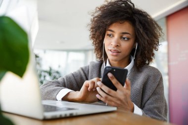 Kafenin penceresinde otururken dizüstü bilgisayar ve cep telefonu kullanan güzel Afro-Amerikan kadının portresi.