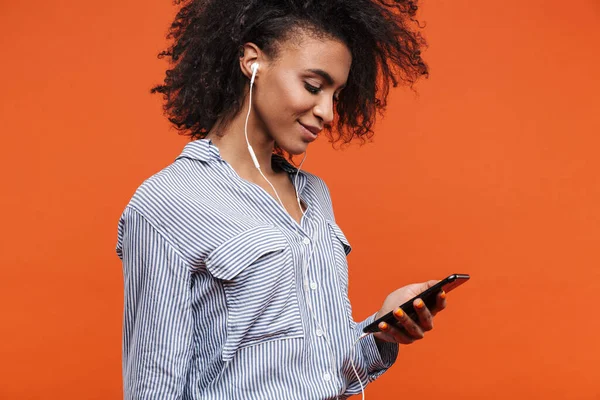 赤い背景に孤立したカジュアルな服を着た若い美しいアフリカ人女性の笑顔 イヤホンや携帯電話で音楽を聴く — ストック写真