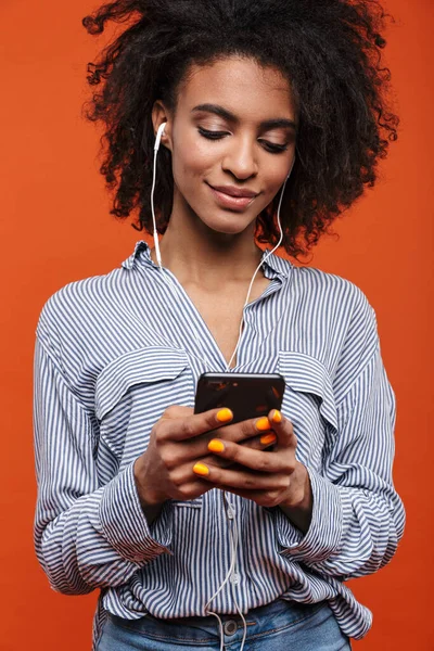 赤い背景に孤立したカジュアルな服を着た若い美しいアフリカ人女性の笑顔 イヤホンや携帯電話で音楽を聴く — ストック写真