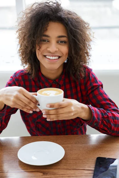 穿着格子呢衬衫微笑着在咖啡馆喝咖啡的漂亮年轻非洲裔美国女人的画像 — 图库照片