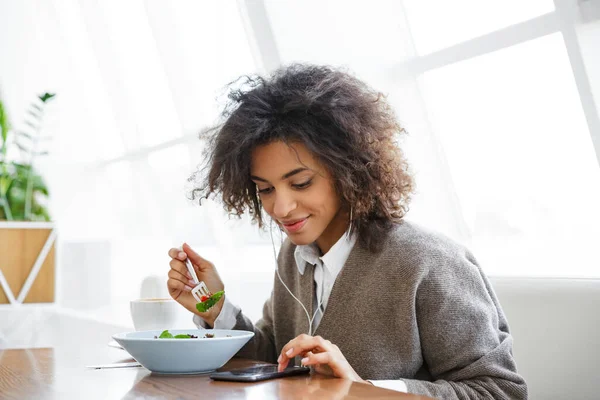 非洲裔美国女青年在咖啡馆里用耳机和手机听音乐的画像 — 图库照片