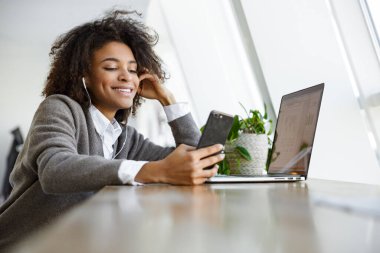 Kafenin penceresinde otururken dizüstü bilgisayar ve cep telefonu kullanan güzel Afro-Amerikan kadının portresi.