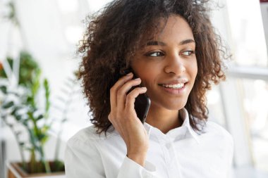Parlak odada gülümseyen ve cep telefonuyla konuşan güzel Afro-Amerikan kadının portresi.