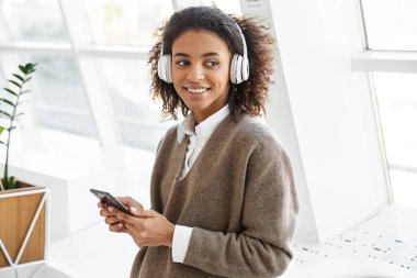 Güzel bir Afro-Amerikan kadının portresi. Parlak odada kulaklık ve cep telefonuyla müzik dinliyor.